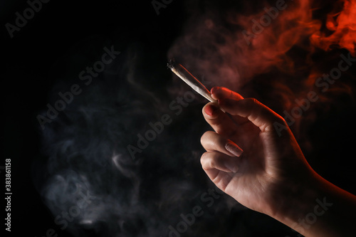 Woman hand holding cigarette in a smoke against black background. Smoking cannabis joint. Medical use. 