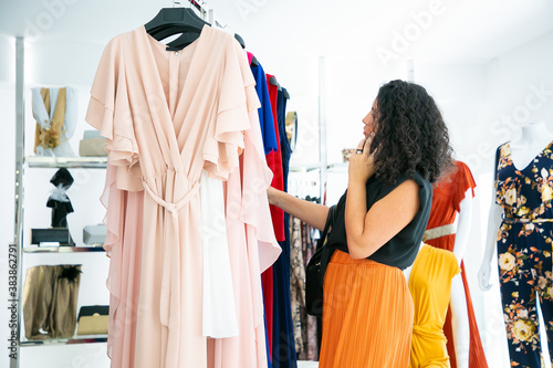 Woman talking on cell while choosing clothes and browsing dresses on rack in fashion store. Medium shot, side view. Boutique customer or retail concept