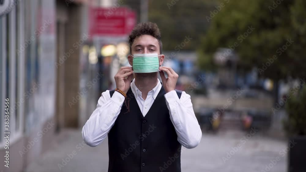 An elegant young man wearing a black waistcoat and a white shirt putting on a green protective Covid-19 mask, standing on a busy street, looking straight into the camera, static close up 4k shot. An elegant young man wearing a black waistcoat and a white shirt putting on a green protective Covid-19 mask, standing on a busy street, looking straight into the camera, static close up 4k shot.