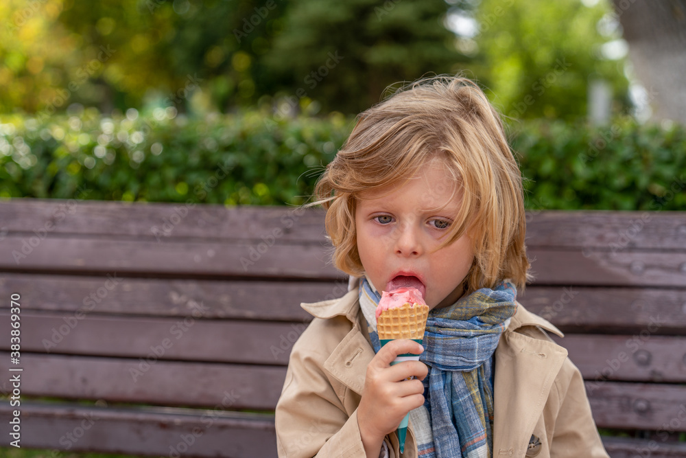 blonde boy in a coat and a scarf eating ice cream while sitting in the park on a wooden bench