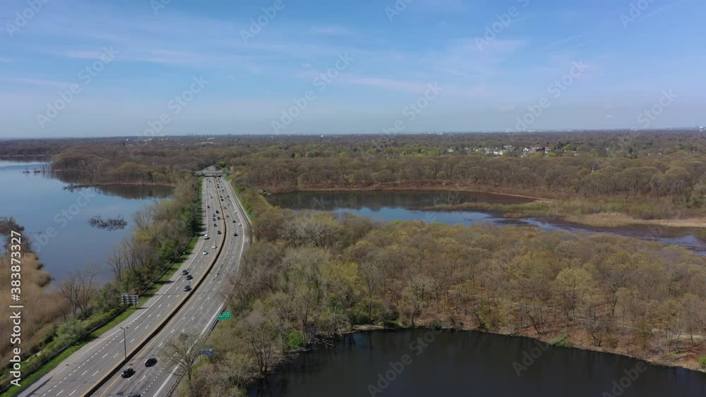 An aerial view of reflective lakes during a clear day. The drone camera dolly out over one of the lake, along a parkway. The parkway below to the left of the shot has car traffic that is moderate. An aerial view of reflective lakes during a clear day. The drone camera dolly out over one of the lake, along a parkway. The parkway below to the left of the shot has car traffic that is moderate.
