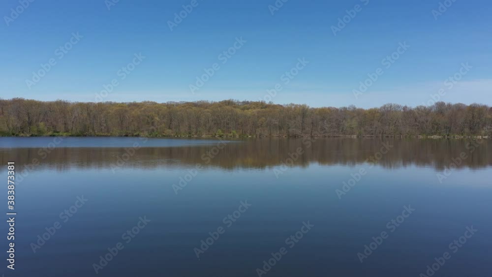 An aerial view of a blue, reflective lake with bare trees lining the lake's edge. The sky is blue & clear. The drone seem to hover as it dolly in slowly towards the trees reflecting onto the waters. An aerial view of a blue, reflective lake with bare trees lining the lake's edge. The sky is blue & clear. The drone seem to hover as it dolly in slowly towards the trees reflecting onto the waters.