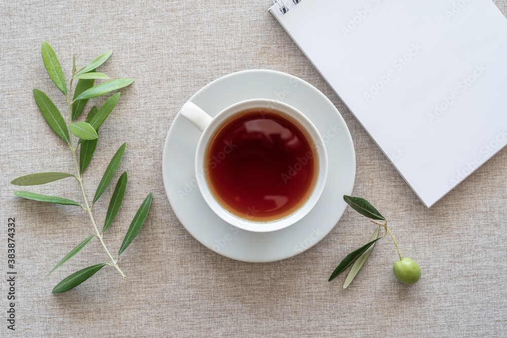 Tea break concept. Tea cup, blank notebook paper, olive branch on cotton fabric background. Tea break concept. Tea cup, blank notebook paper, olive branch on cotton fabric background.