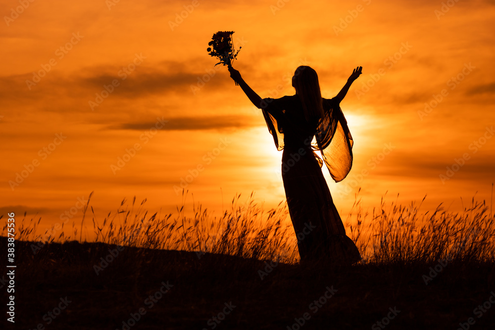 Silhouette of a woman with arms outstretched holding flowers and  looking at beautiful sunset.