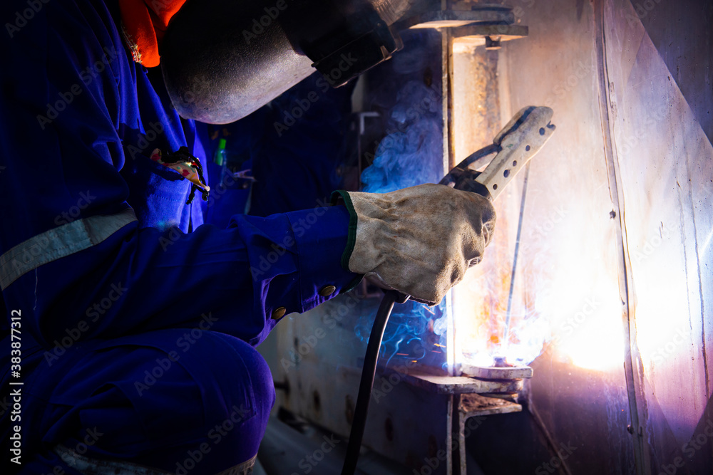 Welder is assembling the workpiece by process shielded metal arc welding (SMAW). Welder in blue uniform, safety shoes, leather gloves, welding mask. He is sitting and welding. Welder is assembling the workpiece by process shielded metal arc welding (SMAW). Welder in blue uniform, safety shoes, leather gloves, welding mask. He is sitting and welding.