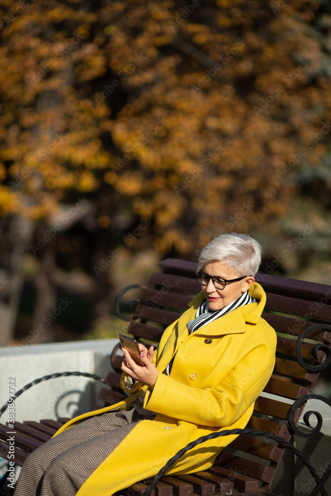 an adult woman in age sits on a bench with a mobile phone in her hands in a park in autumn in a yellow coat