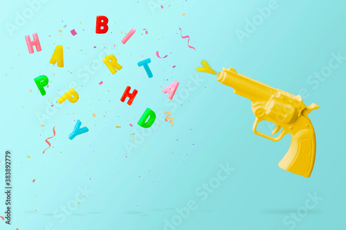 Yellow pistol shooting colorful birthday candles and colorful confetti, against blue background. Happy birthday concept with copy space