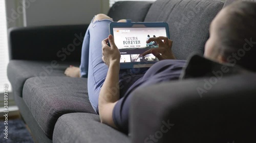 Woman using movie stream service with tablet. Choosing series or film and lying on couch in home living room. Watching an episode of a tv show with mobile device. Streaming on demand video.