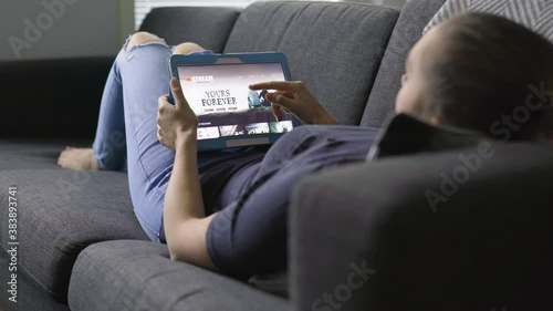 Movie stream service online. Woman choosing series or film with tablet and lying on couch in home living room. Streaming on demand video or watching an episode of a tv show with mobile device.