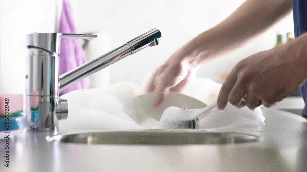 Man washing dishes in the kitchen sink. Person doing domestic household chores. Turn off the water from tap and start to scrub white plate with brush. Soap foam and bubbles. Man washing dishes in the kitchen sink. Person doing domestic household chores. Turn off the water from tap and start to scrub white plate with brush. Soap foam and bubbles.