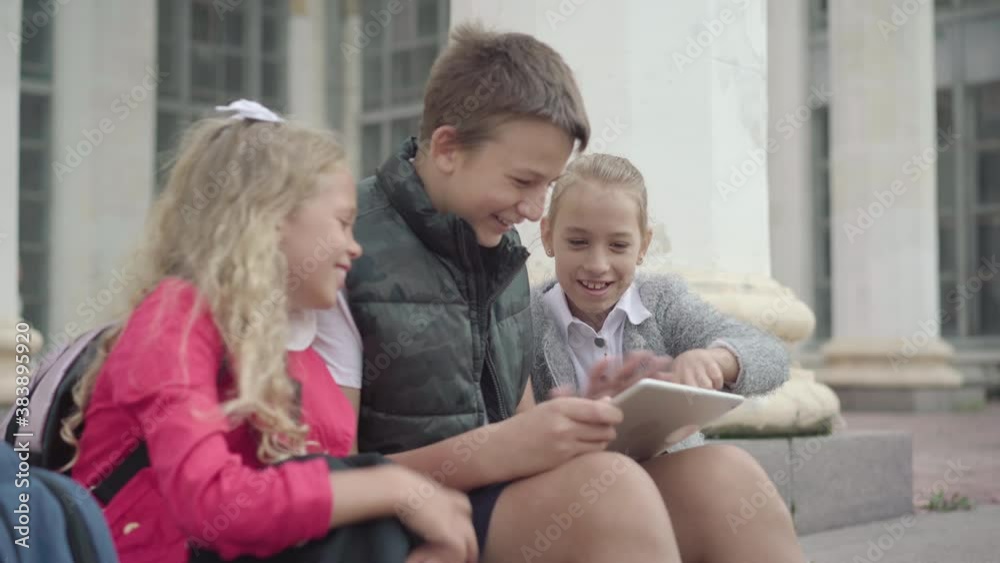 Three cheerful children classmates using social media on tablet and talking. Side view portrait of happy Caucasian schoolboy and schoolgirls laughing and chatting on break. Three cheerful children classmates using social media on tablet and talking. Side view portrait of happy Caucasian schoolboy and schoolgirls laughing and chatting on break.