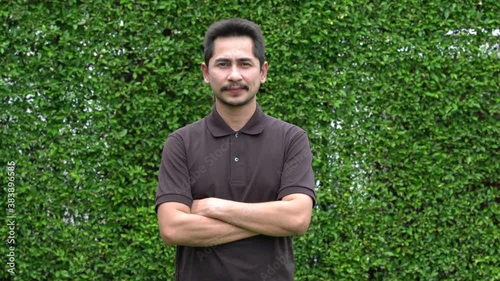 Young man asian smiling and looking at camera. Portrait of a happy handsome young man on green nature background. Young cool trendy cross one's arm with confident. Young man asian smiling and looking at camera. Portrait of a happy handsome young man on green nature background. Young cool trendy cross one's arm with confident.