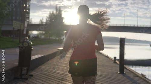Person running in city. Woman on a morning jog. Following slow motion back view. Fitness runner training. Sunny sky with clouds at sunset or sunrise. Jogger on dock by the lake. Sport motivation.