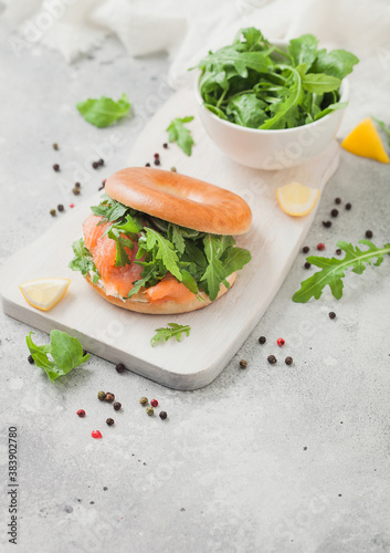 Bagel and salmon sandwich with cream cheese and wild rocket in white bowl and lemon with pepper on light background.