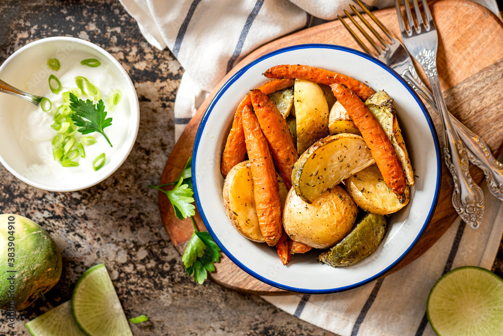 Baked vegetables. Roasted potatoes, carrots and radish with herbs in a bowl on a dark background top view. Vegetarian food, rustic style.