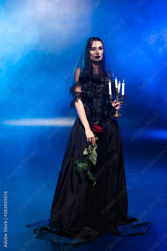 bride in black dress and veil holding rose and candles on blue with smoke, halloween concept