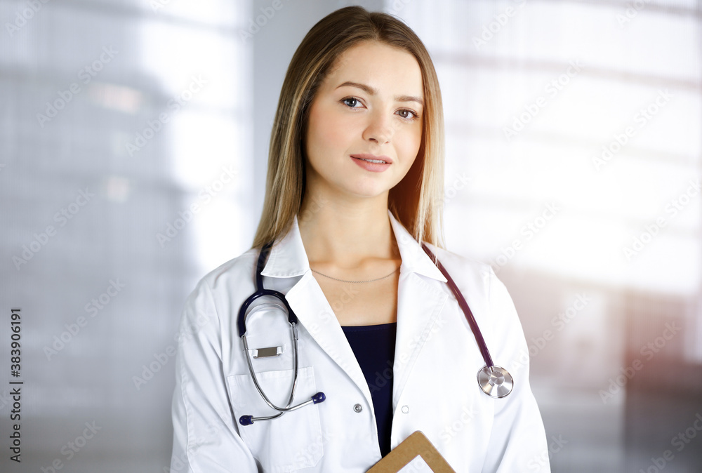 Young optimistic woman-doctor is holding a clipboard in her hands, while standing in a sunny cabinet of a clinic. Portrait of friendly female physician with a stethoscope. Perfect medical service in a Young optimistic woman-doctor is holding a clipboard in her hands, while standing in a sunny cabinet of a clinic. Portrait of friendly female physician with a stethoscope. Perfect medical service in a