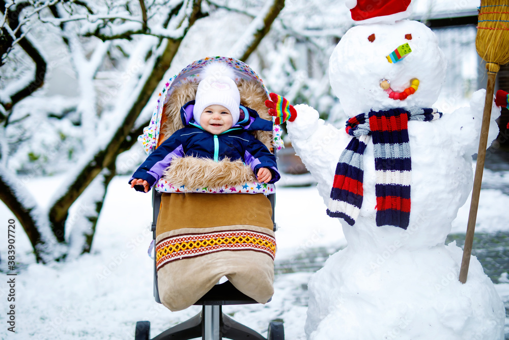 Cute little beautiful baby girl sitting in the pram or stroller on winter day with snow and snowman