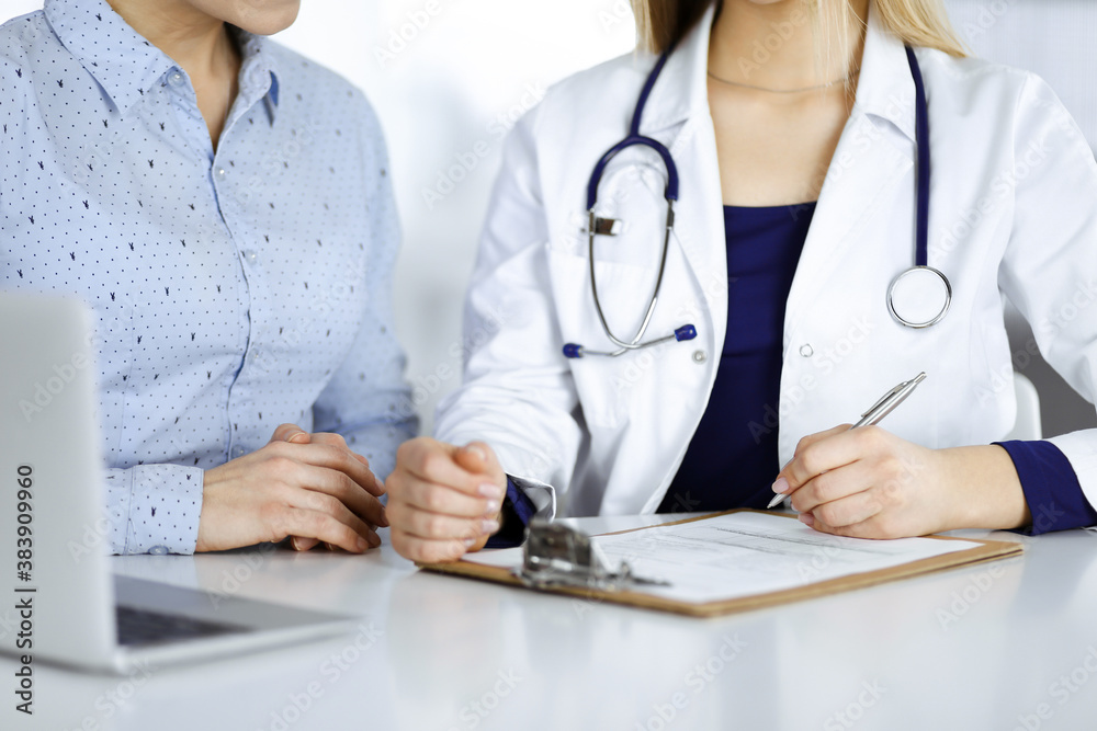 Unknown woman- doctor and her patient are discussing patient's blood test, while sitting together at the desk in the cabinet in a clinic. Female physician, with a stethoscope, is using a laptop Unknown woman- doctor and her patient are discussing patient's blood test, while sitting together at the desk in the cabinet in a clinic. Female physician, with a stethoscope, is using a laptop