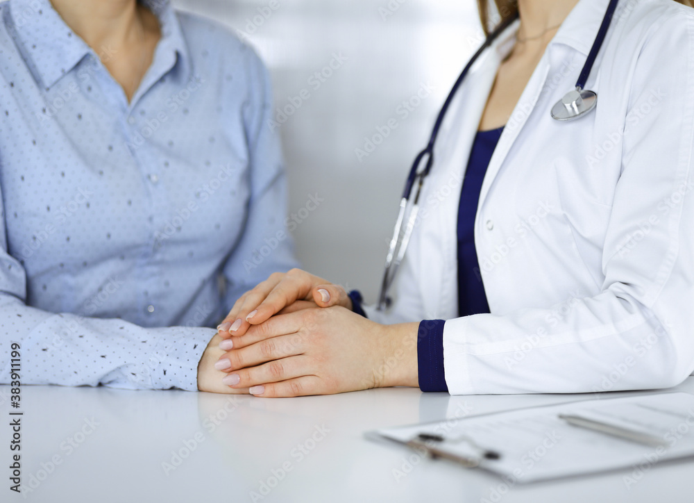 Unknown woman-doctor is holding her patient's hands to reassure a patient, while sitting together at the desk in the cabinet in a clinic. Female physician is using a clipboard and a stethoscope, close Unknown woman-doctor is holding her patient's hands to reassure a patient, while sitting together at the desk in the cabinet in a clinic. Female physician is using a clipboard and a stethoscope, close