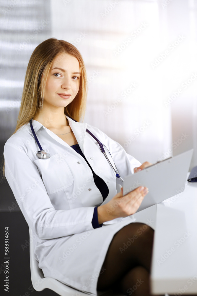 Young optimistic woman-doctor is using a clipboard, while sitting at the desk in her sunny cabinet in a clinic. Portrait of friendly female physician woman with a stethoscope. Perfect medical service Young optimistic woman-doctor is using a clipboard, while sitting at the desk in her sunny cabinet in a clinic. Portrait of friendly female physician woman with a stethoscope. Perfect medical service