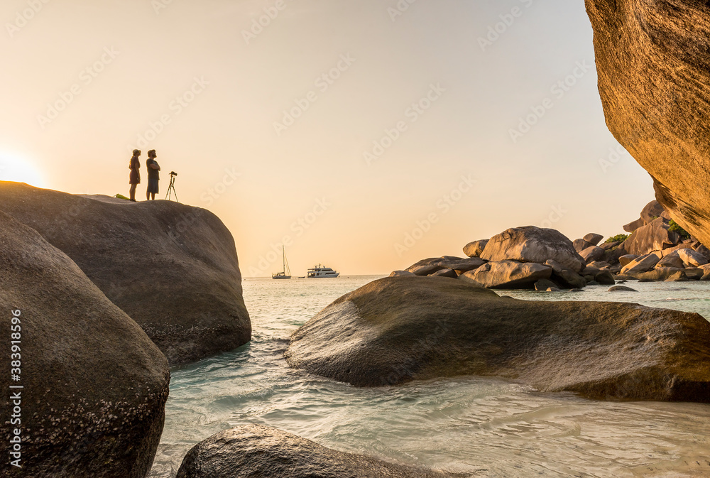 A girl and a man photographer shoot the tourist ship in the sea at sunset in Thailand standing on a huge rock