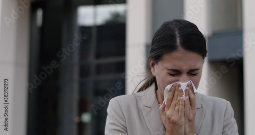 Close up of sick young female standing on street and coughing and sneezing. Portrait of unwell beautiful Caucasian woman blowing runny nose into tissue outdoors. Infection. Virus and disease concept photo