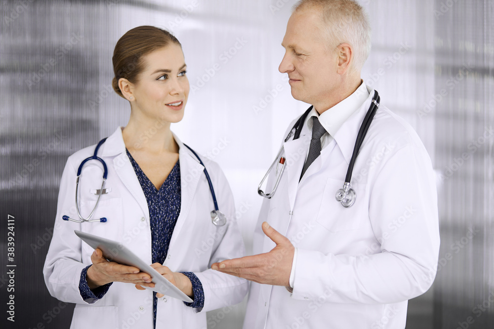 Fototapeta premium Elderly confident doctor and woman physician discussing current disease therapy while standing in clinic. Teamwork in hospital. Medicine and healthcare concept