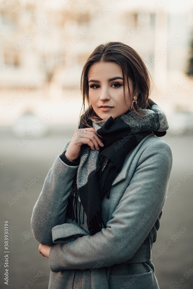 Vertical portrait of young indian woman dressed in grey coat with glasses and scarf standing on the street, looking at the camera, image with copy space and blur background