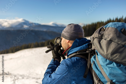 Hiker man tourist photographer in warm clothing with backpack and camera taking picture of snowy valley and woody mountain peaks landscape under blue sky on sunny winter cold day. © bilanol