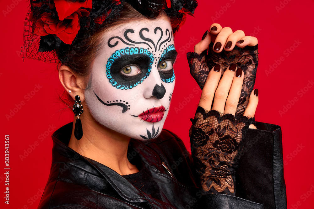 Sugar skull pretty girl with halloween makeup standing on bright red background. Wonderful female zombie with flowers in hair celebrating day of the dead.