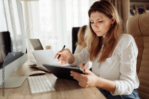 Confused woman sit at the table, look at the tablet, having problems with gadget or internet connection, misunderstanding with colleagues, young mother working from home online