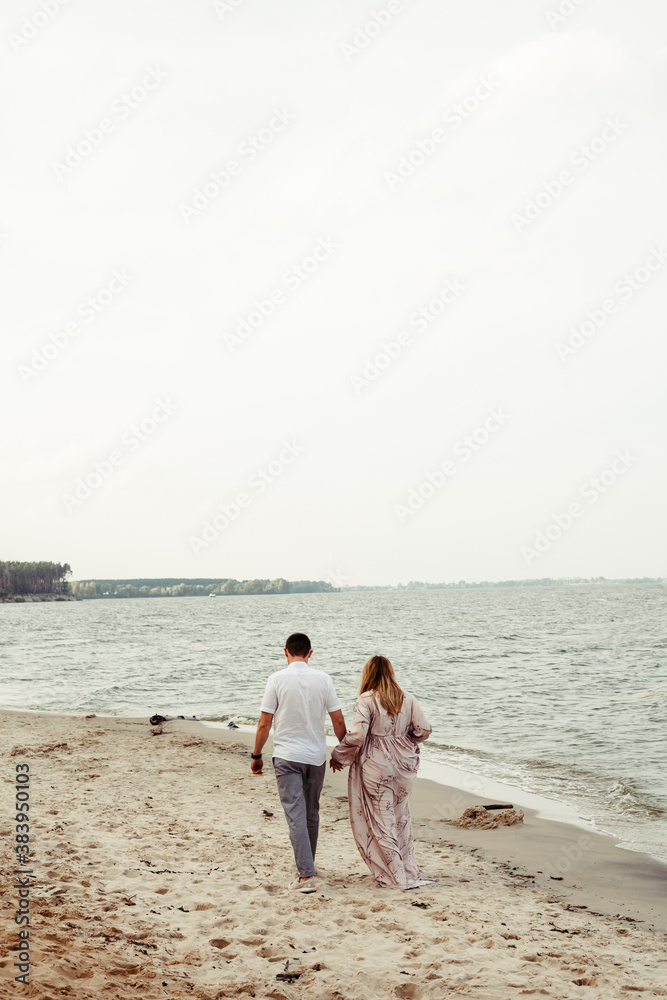 A pregnant woman and her husband are walking by the hand against the background of the sea. Place for an inscription. Couple in love
