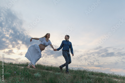 A couple in love, a pregnant wife in a white outfit are walking in a hilly field. Overcast weather