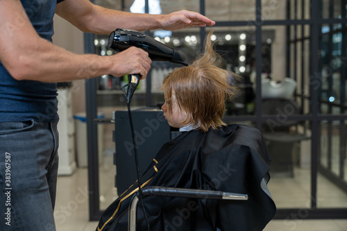 in a beauty studio, a stylist makes a styling to a blonde boy using a hairdryer
