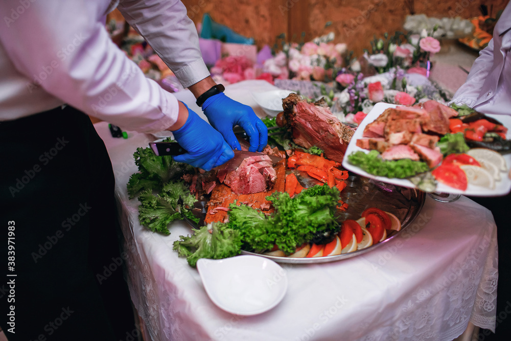 Male chef cuts meat with a knife in a restaurant, a large piece of cooked meat dish on a tray with herbs and vegetables, the waiter serves food to the guests. Free space for text