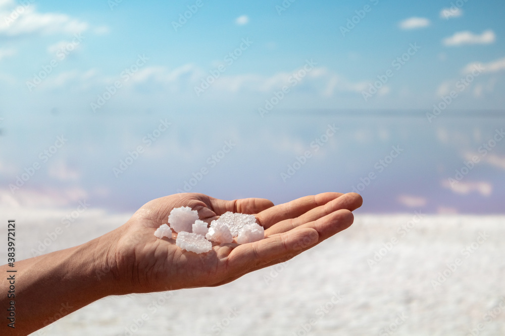 Hand holding bunch of pink white salt flakes crystals with pink salty lake and blue sky background. Spa resort sunny close-up Hand holding bunch of pink white salt flakes crystals with pink salty lake and blue sky background. Spa resort sunny close-up