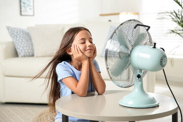Little girl enjoying air flow from fan at home. Summer heat