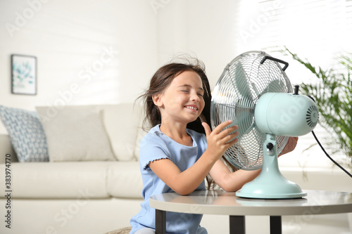 Little girl enjoying air flow from fan at home. Summer heat photo