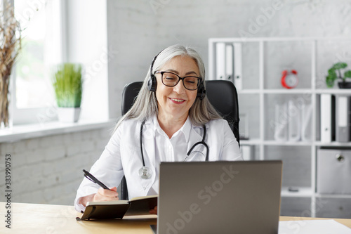 Portrait of senior grey-haired female doctor in her office using laptop for video chat with a patient. Online consultation with doctor for diagnoses and treatment recommendation. Telehealth concept. photo