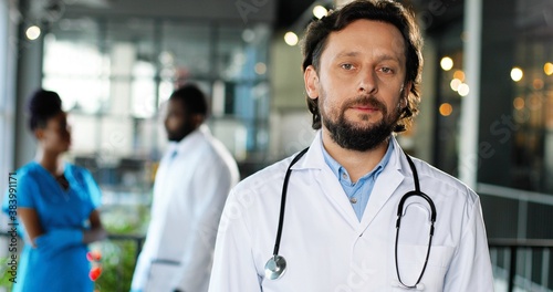 Portrait of Caucasian man physician in white gown looking at camera, smiling and standing in clinic. Handsome male doctor in hospital. African American doctors on background. Medicine concept.