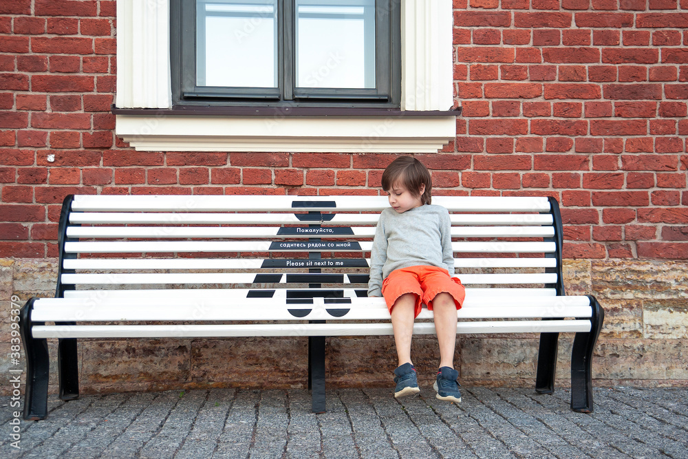 The boy sits on the bench and looks at the sign of social distance.