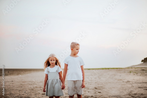 A beautiful little brother in a white t-shirt holding hand blonde younger sister 