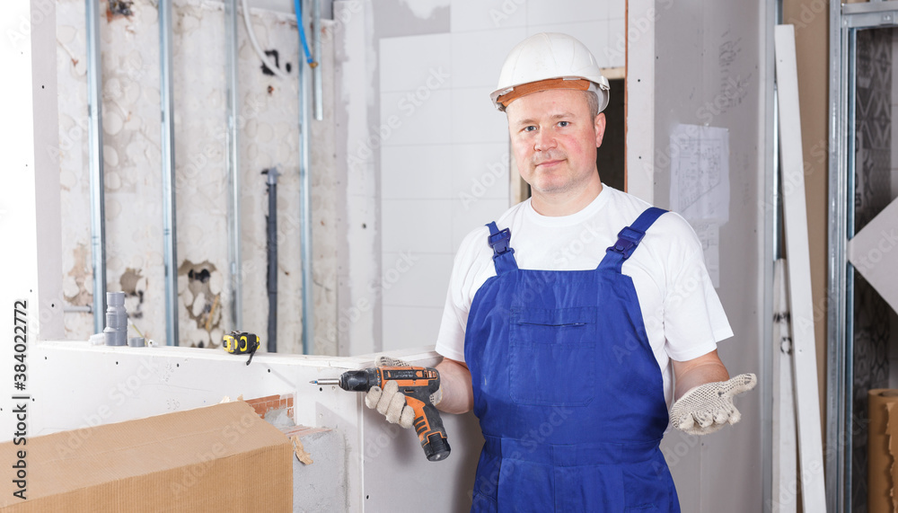 Confident male contractor engaged in drywall mounting using screw gun in overhauls indoors.