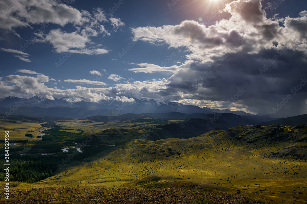 Obraz premium Majestic panorama of mountain plain on the background of snow-covered ridge before thunderstorm. Sun's rays break through huge clouds and beautifully illuminate green steppe and winding river.