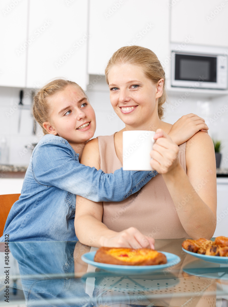 Cheerful teenage girl and her mother sitting at kitchen table and drinking tea