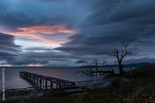 View of a pier and skeletal trees on a lake at dusk  beneath a dramatic  moody sky