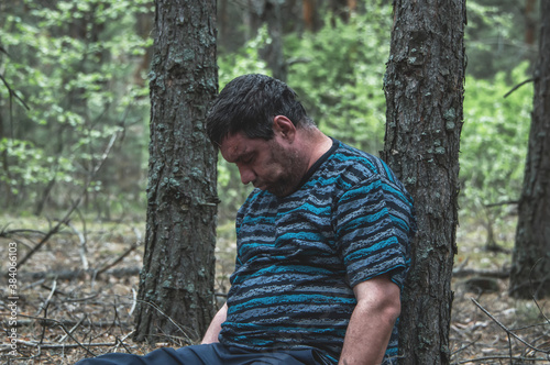Murder in the woods. The body of a man in a blue t-shirt and trousers is sitting on the ground among the trees in the forest. Victim of an attack. Horizontal photo.