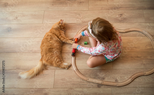 Portrait of cute happy caucasian kid girl and red kitten. Little positive kid sitting and playing on the floor with wooden train road. Warm floor for child and domestic animal.