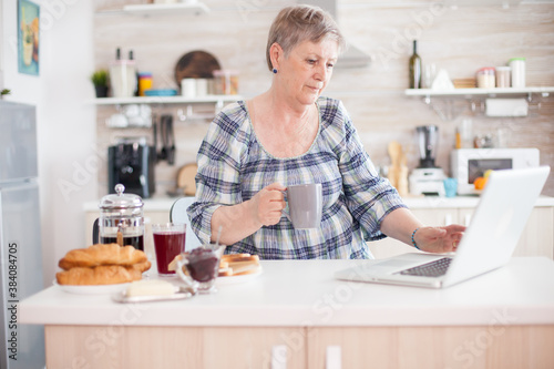 Senior woman using laptop in kitchen during breakfast and driking coffee. Elderly retired person working from home, telecommuting using remote internet job online communication on modern technology